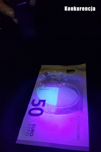 Banknot 50 euro podświetlony światłem UV w ciemnym otoczeniu, pokazujący zabezpieczenia antyfałszywkowe. Na górze napis: „Konkurencja”.
