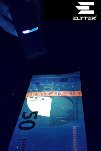 Lampa UV oświetla banknot 50 euro w ciemnym otoczeniu, ukazując zabezpieczenia antyfałszywkowe; logo Elyter widoczne.