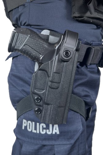 Pistolet w kaburze przypiętej do pasa policjanta w granatowym mundurze z napisem "POLICJA" na spodniach, sprzęt taktyczny.